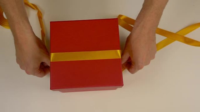 准备礼品包装。一盒红色的纸板。男士手测量金缎丝带来装饰礼品盒。俯视图特写。黄色胶带。