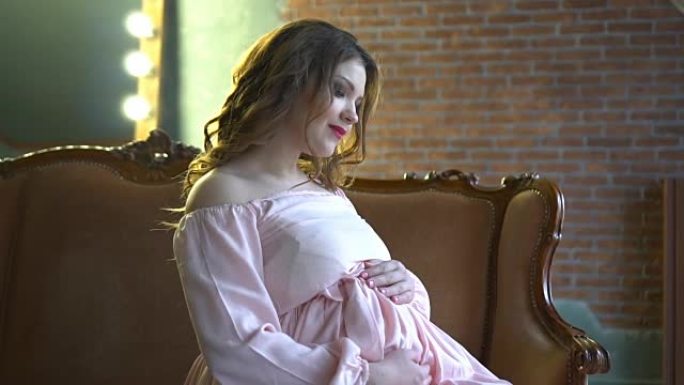 穿着优雅粉色连衣裙化妆的美丽孕妇坐在家里的沙发上