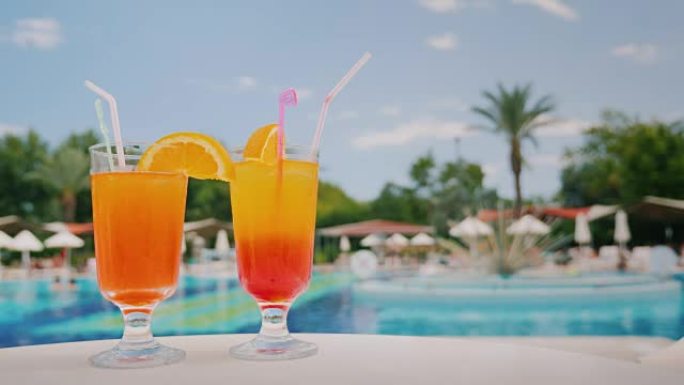 两杯凉爽的鸡尾酒站在游泳池和棕榈树背景的桌子上。天堂和豪华度假村