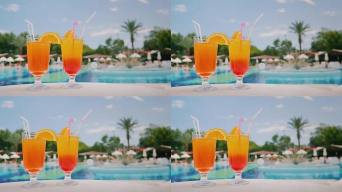 两杯凉爽的鸡尾酒站在游泳池和棕榈树背景的桌子上。天堂和豪华度假村