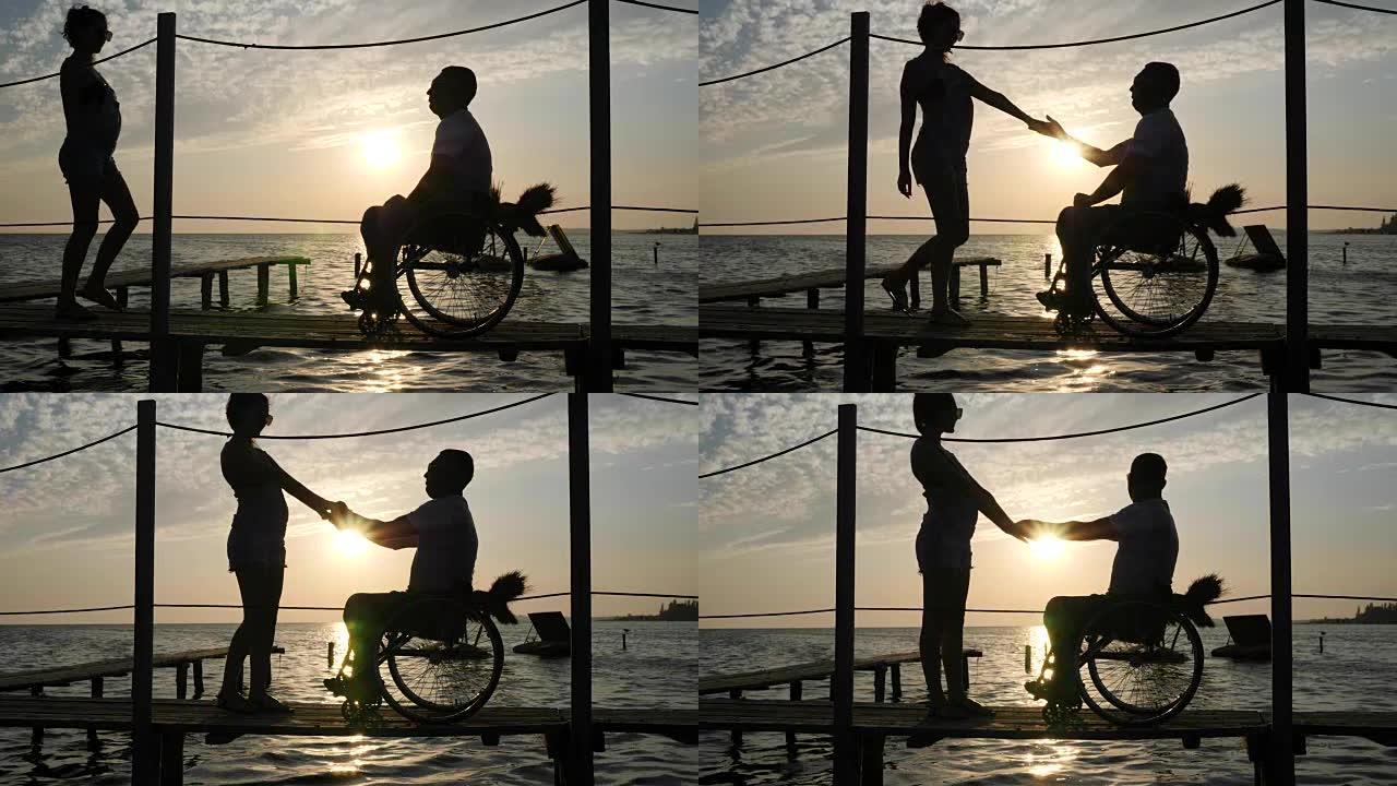 在明亮的光束中闪亮的水附近的海滨与残疾人的情侣的剪影