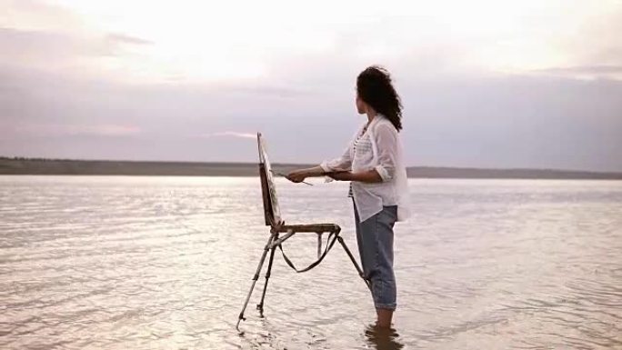 一个年轻卷曲的黑发女人站在水中，用画架吸引她周围的风景。穿着蓝色牛仔裤和白色衬衫。全长