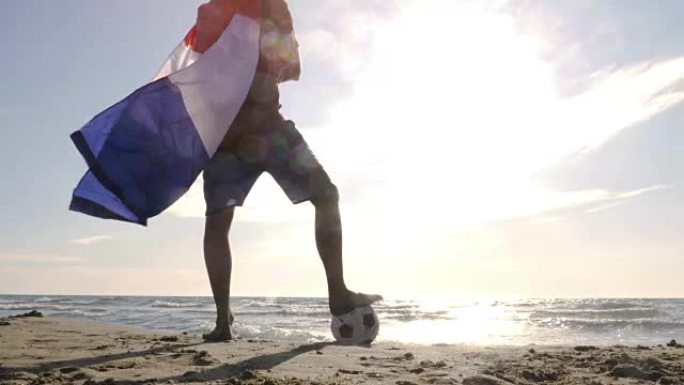 戴着法国国旗的年轻人作为超级英雄斗篷，脚踩着足球站在海边的沙滩上，日落时看着海滩上的海洋摄像机ste