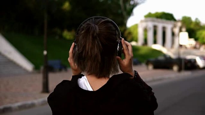 一个穿着黑色休闲衣服的年轻现代女孩在公园或街道上散步的背面视图。回头看她的肩膀，微笑着。戴上无器皿耳