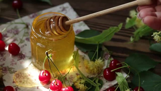 打开玻璃罐液体蜂蜜和蜂蜜北斗七星，一束椴树花和红樱桃在木制表面上。一缕阳光。深色乡村风格。