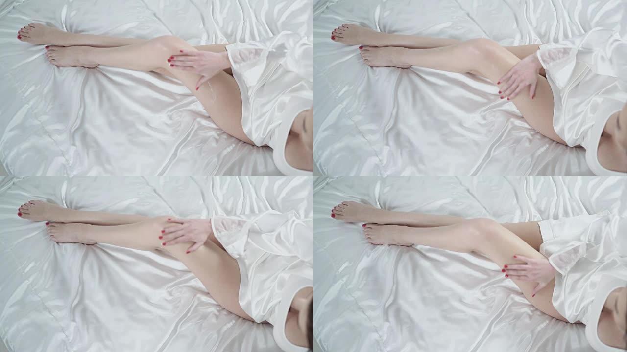 美女坐在床上，在光滑的腿上涂上奶油。腿部皮肤护理。