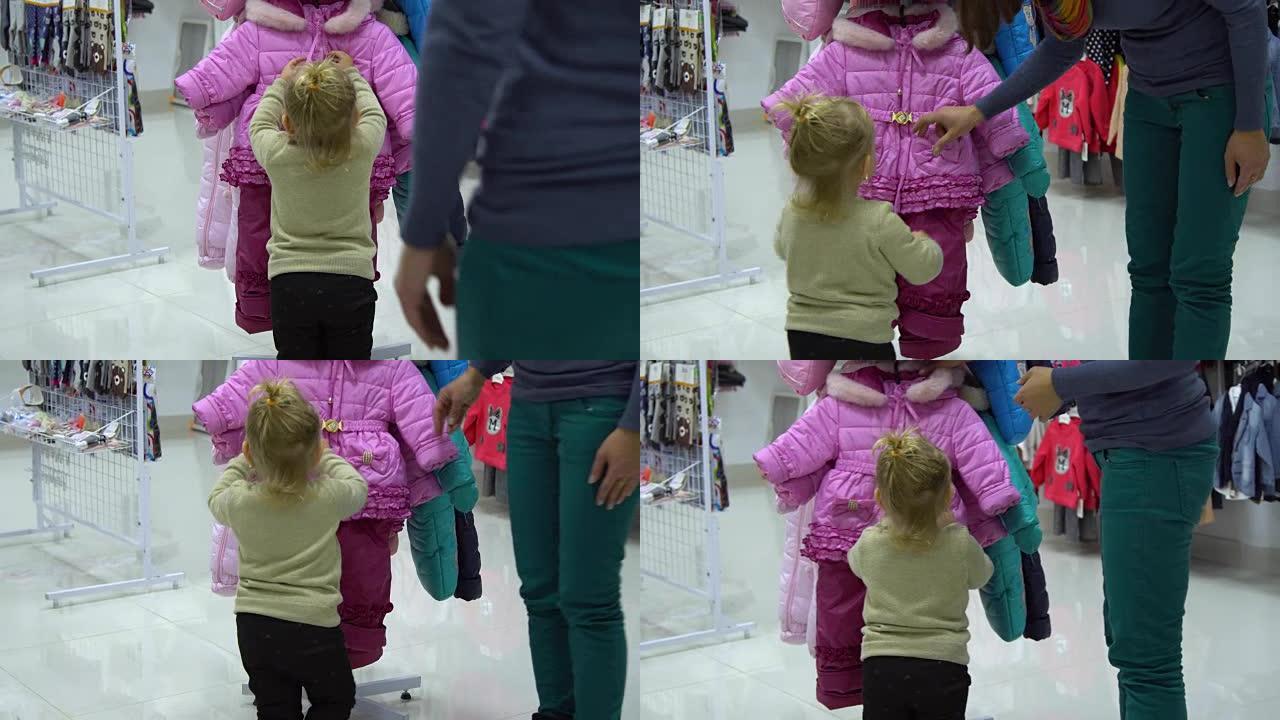 一个可爱的小女孩和她的母亲在精品店里选择童装。妈妈和孩子在商店里挑选衣服。考虑一套儿童冬季套装。
