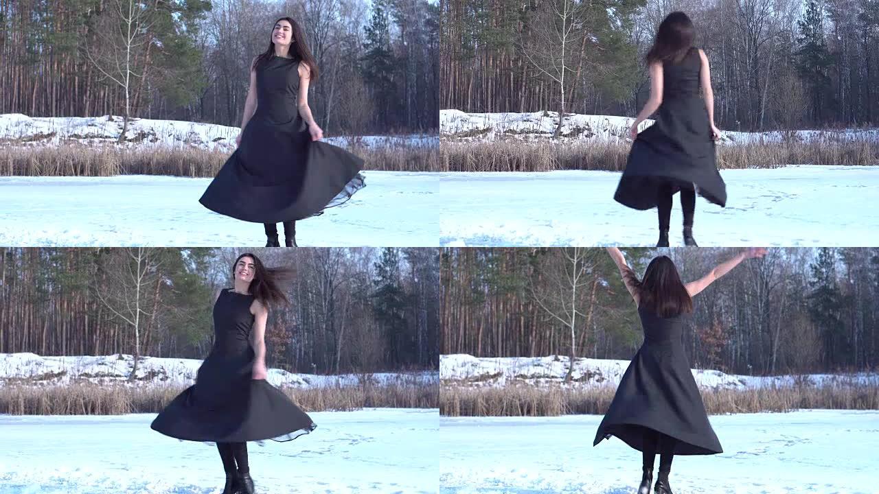 年轻微笑的黑发女孩穿着黑色连衣裙转过身来，把手举在冬季森林景观背景上