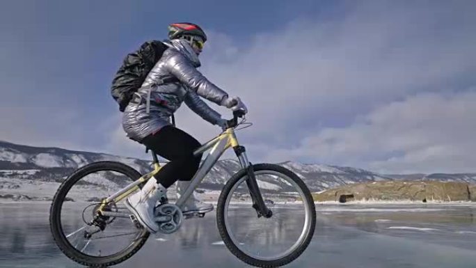女人在冰上骑自行车。这个女孩穿着银色羽绒服，骑自行车背包和头盔。冰冻的贝加尔湖的冰。自行车上的轮胎上
