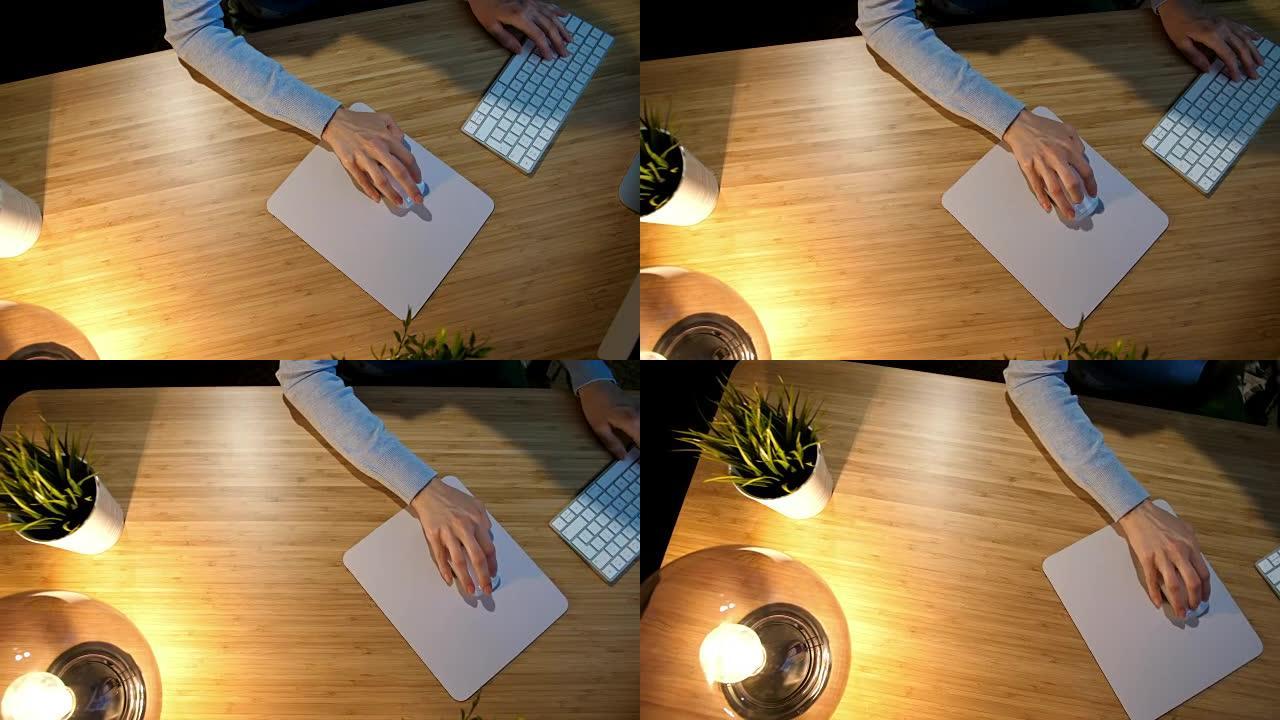 晚上在电脑上工作的女性。穿着舒适的家居服的女人的手的裁剪视图，晚上坐在灯点亮的木桌上，在黑暗的房间里