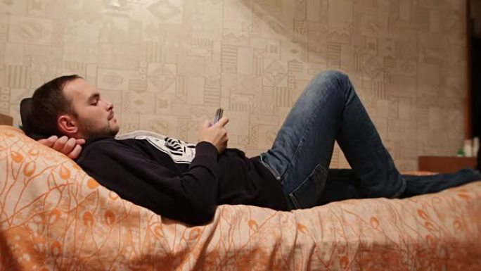 留着胡子的放松懒人躺在家里的床上，在肖像模式下使用smatphone。懒惰的高加索人在手机上看新闻或