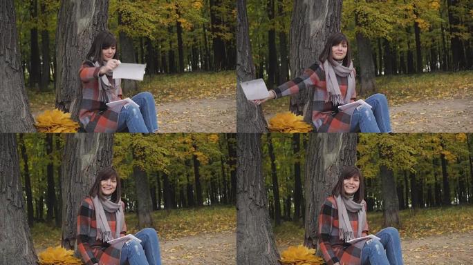 从笔记本上拿一片叶子。迷人的年轻快乐微笑的女人坐在公园的草地上，在笔记本上做笔记。