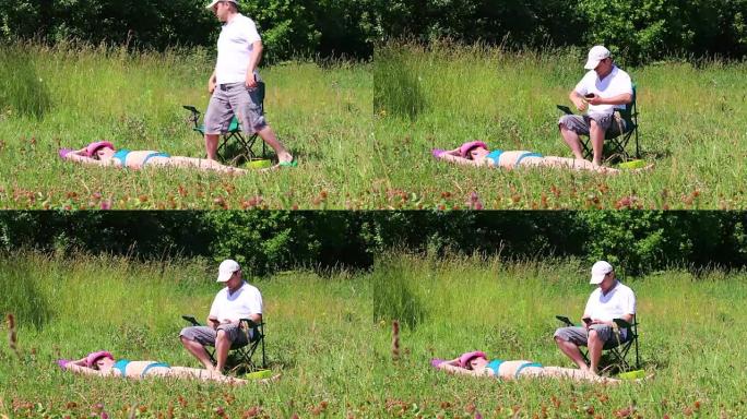 一个穿着泳衣的女孩正在草坪上晒日光浴。在她旁边的野餐椅上，一个男人坐在智能手机上玩耍。