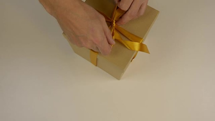 特写俯视图高角度。男人的手在棕色纸箱礼品盒上用缎带做蝴蝶结。白色米色背景。准备庆祝节日的礼物。圣诞礼