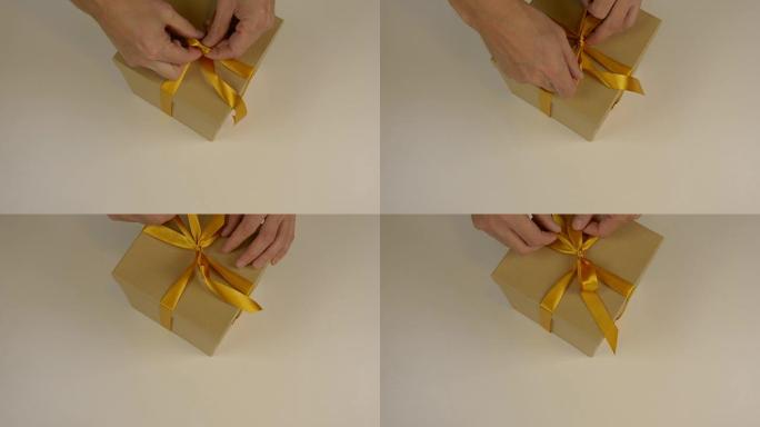 特写俯视图高角度。男人的手在棕色纸箱礼品盒上用缎带做蝴蝶结。白色米色背景。准备庆祝节日的礼物。圣诞礼