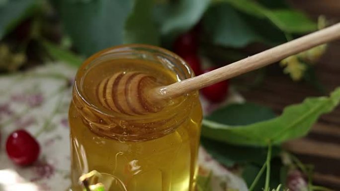 打开玻璃罐液体蜂蜜和蜂蜜北斗七星，一束椴树花和红樱桃在木制表面上。一缕阳光。深色乡村风格。