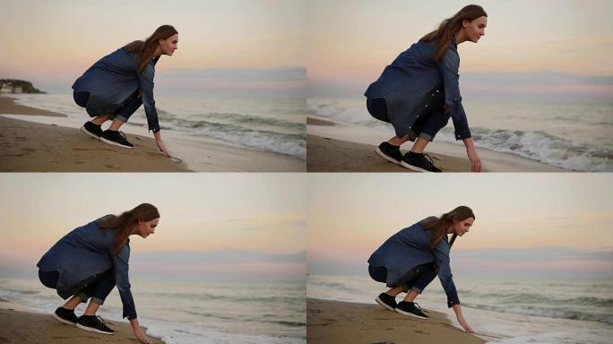 迷人的年轻女子坐在海边，在日落时用手在海滩上尝试水温。慢动作镜头