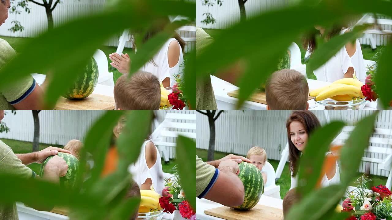 夏天，一个年轻的家庭，有两个有趣的小孩坐在花园里的餐桌旁。爸爸切了一个大西瓜，要请他的家人。与家人共