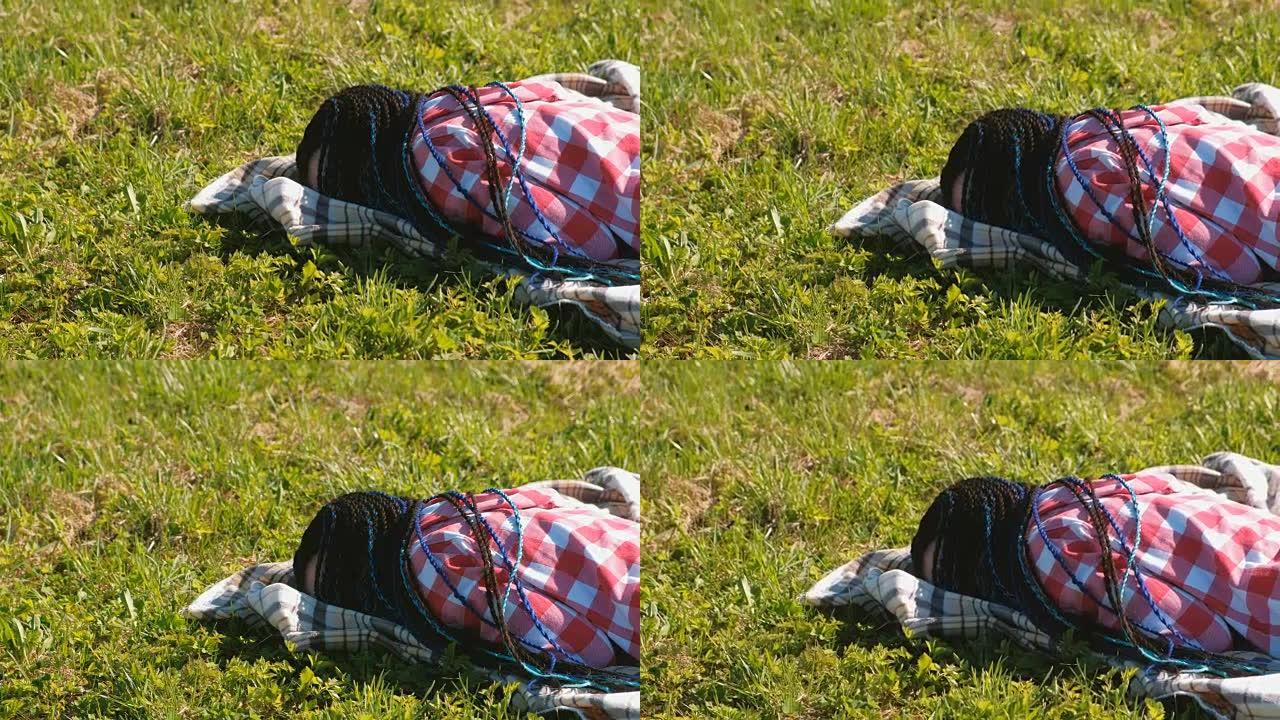 无法辨认的蓝色非洲辫子的女人睡在公园的草坪上。
