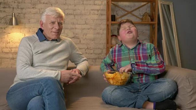 祖父和他的孙子坐在沙发上看电视，吃薯条，微笑着。一位老人切换频道，使用遥控器。家庭舒适，家庭田园诗，