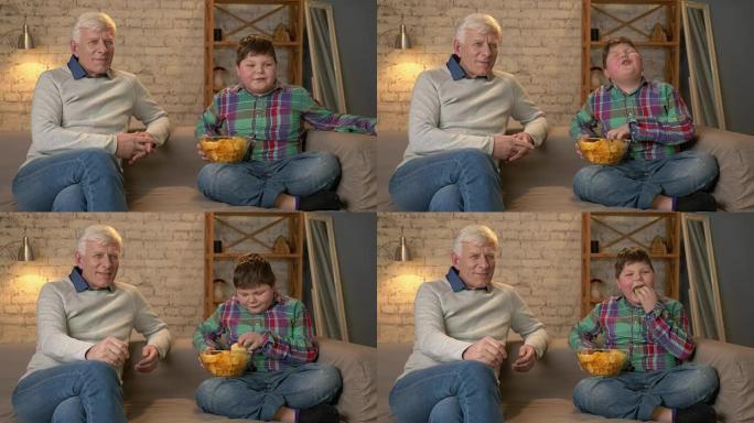 祖父和他的孙子坐在沙发上看电视，吃薯条，微笑着。一位老人切换频道，使用遥控器。家庭舒适，家庭田园诗，