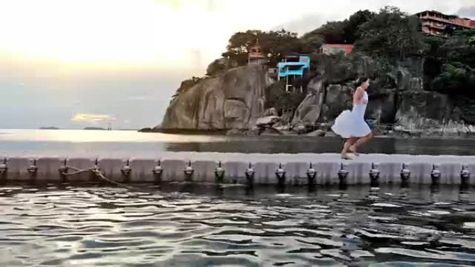 在美丽的日落时分，穿着白色连衣裙的年轻黑发女子在热带岛屿大海中的桥上奔跑。1920x1080