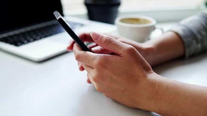 坐在咖啡馆里，桌上放着一杯咖啡和笔记本电脑，在智能手机上使用应用程序的女性手的特写视图
