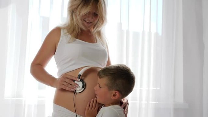 男孩听着母亲怀孕的肚子，女人拿着耳机在裸露的大肚子上微笑