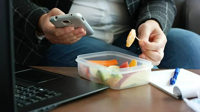 节食西装的女商人从午餐盒里吃了健康的水果零食，看着智能手机