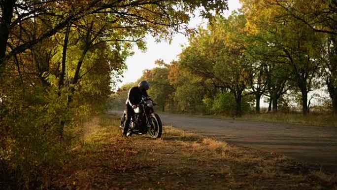 穿着头盔和皮夹克的面目全非的时尚男人在秋天的阳光明媚的日子里站在路边时骑着自行车启动引擎