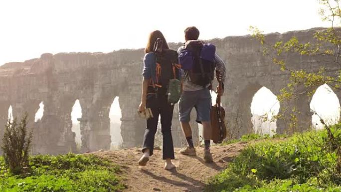 年轻可爱的夫妇背包客游客手牵着手走向罗马渡槽拱门，在浪漫的迷雾日出时，带着吉他和睡袋慢动作