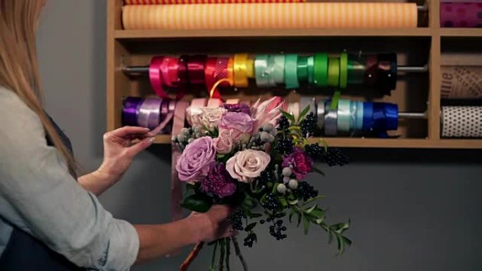 女性金发花店安排现代花束，并为它选择完美的riband结合不同的颜色的后视图。英俊的花店老板在她的花