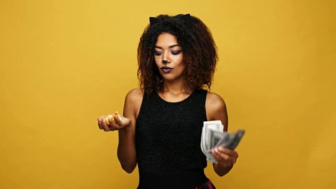 无忧无虑的非洲女士与猫化妆手握糖果和金钱，捣蛋和选择金钱隔离在黄色