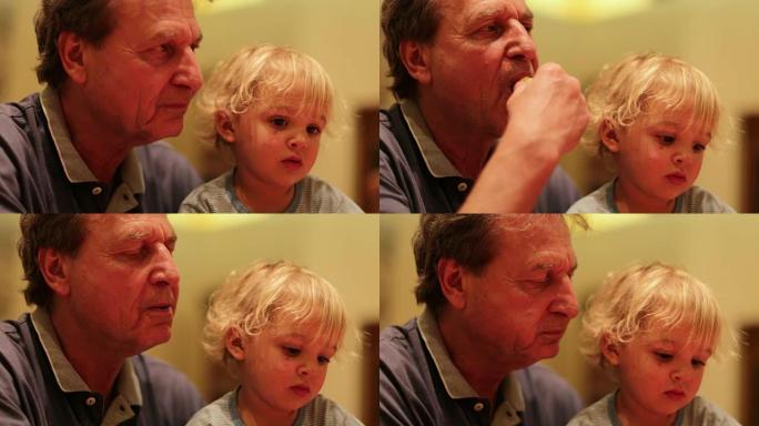 祖父和他的孙子。爷爷吃饭时抱着蹒跚学步的坦率时刻。年长的男人抱着婴儿时咀嚼薯条