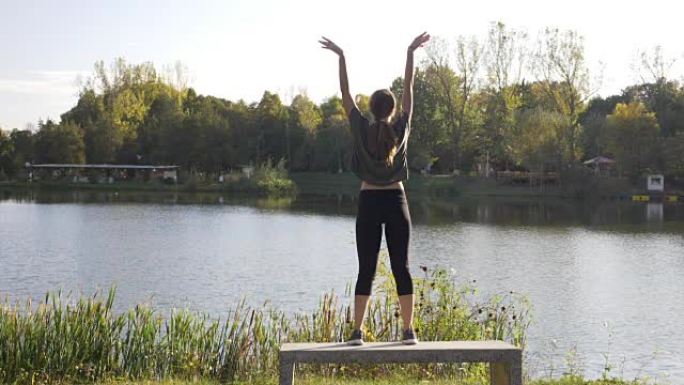 穿着运动服的瘦瘦迷人的女孩在公园户外练习瑜伽，站在湖边的长椅上慢动作