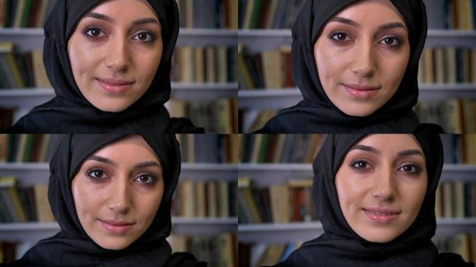 戴着头巾的年轻美丽的穆斯林女孩正在看书，看着相机，宗教概念，背景书架，背景模糊