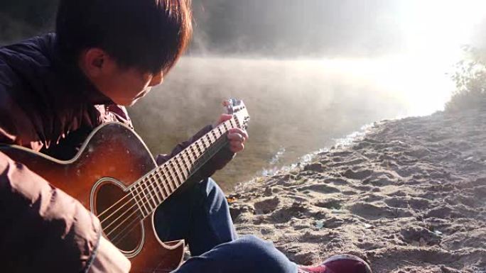 英俊的亚洲年轻人在日出时在森林河海岸弹吉他。背景下的黎明迷雾