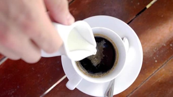 男性手倒牛奶的俯视图在木制表面上的白色咖啡杯中倒入浓缩咖啡。慢动作。3840x2160