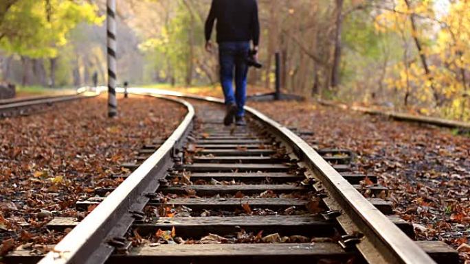 一个拿着DSLR相机的男人在背景的秋天森林中沿着火车轨道走。一名男子离开摄像机