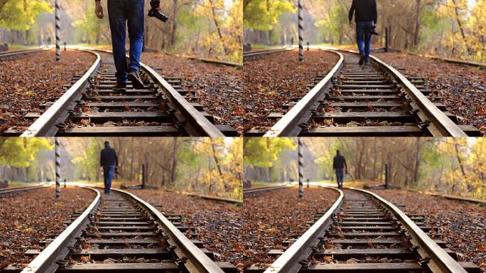 一个拿着DSLR相机的男人在背景的秋天森林中沿着火车轨道走。一名男子离开摄像机