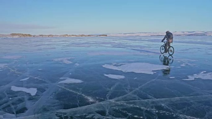 人在冰上骑自行车。骑自行车的人穿着灰色羽绒服，背包和头盔。冰冻的贝加尔湖的冰。自行车上的轮胎上覆盖着