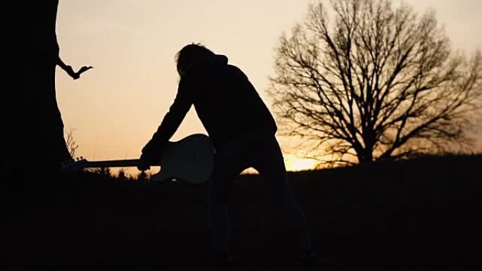 人在日落时在树附近的田野里弹电吉他，唱一首抒情歌。剪影