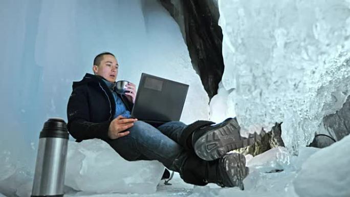 男人坐在互联网上的笔记本电脑在一个冰洞里。在神秘美丽的冰窟周围。用户在社交网络中交流。他穿着黑色外套