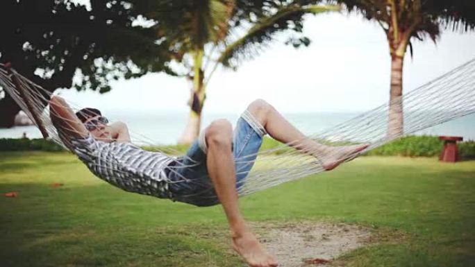 戴着墨镜的年轻人在美丽的热带海滩上的吊床上放松。度假概念。1920x1080