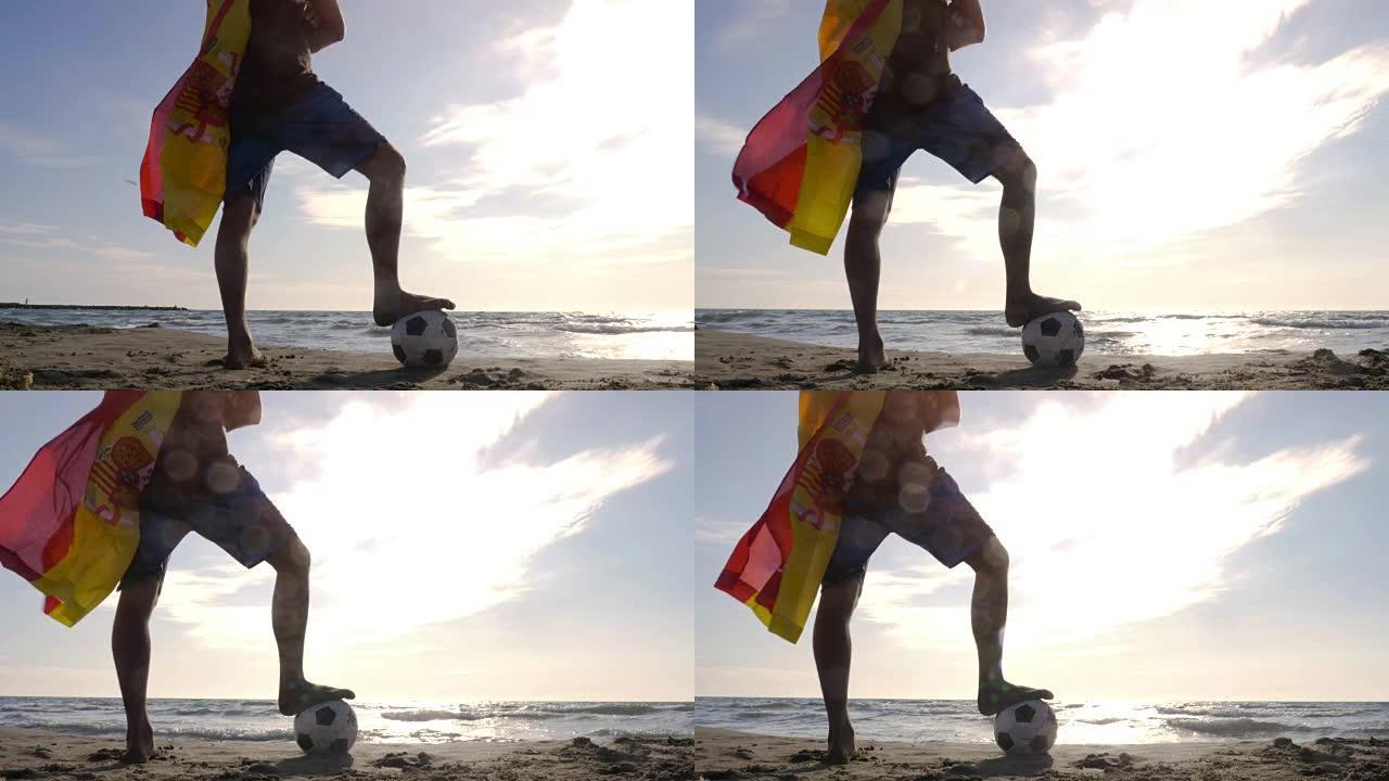 穿着西班牙西班牙国旗作为超级英雄斗篷的年轻人脚踩着足球站在海边的沙滩上，日落时看着海滩上的海洋摄像机