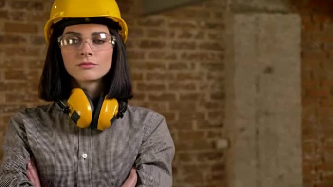 年轻迷人的建筑工人站在耳机、眼镜和头盔里，双手交叉看着相机，砖砌建筑背景