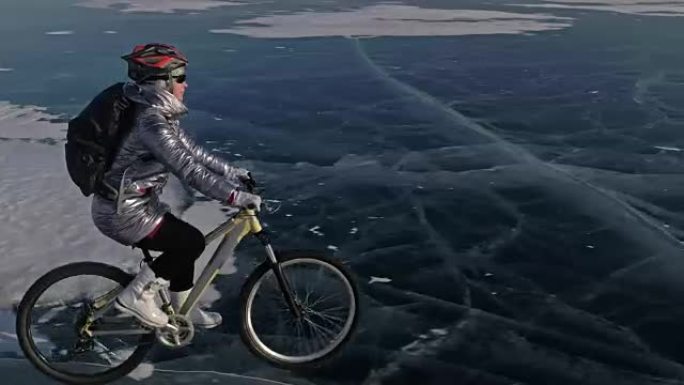 女人在冰上骑自行车。这个女孩穿着银色羽绒服，骑自行车背包和头盔。用四旋翼无人机射击。自行车上的轮胎上