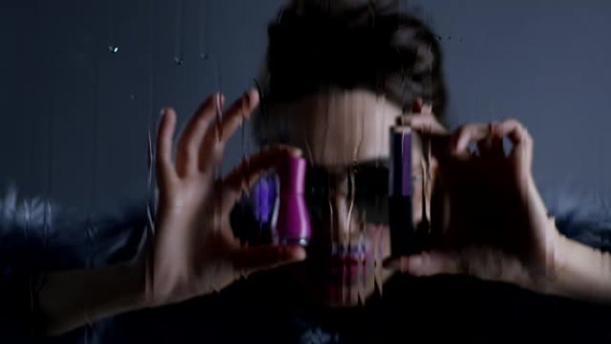 戴着太阳镜和皮草的黑发高级时装模特一只手拿着粉色指甲油，另一只手拿着紫色唇膏。时尚视频。