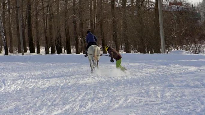 慢动作: 一个女孩骑着马疾驰。一匹马在绳子上拖着一个滑雪者。滑雪者在雪堆中骑在滑雪板上。女骑师和男滑