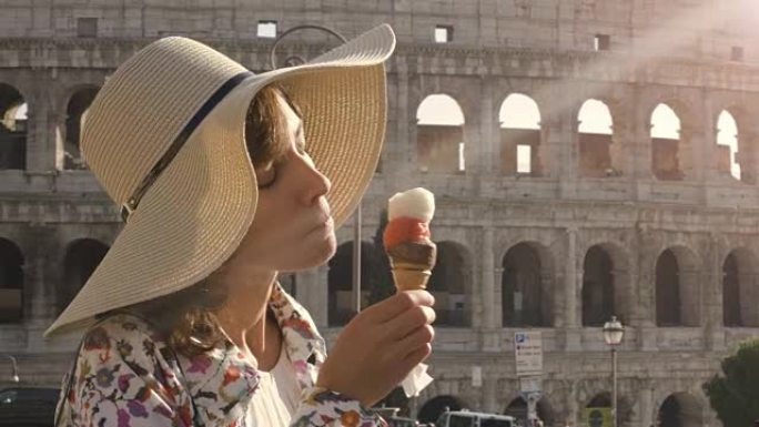 美丽的年轻女子穿着五颜六色的时尚连衣裙在罗马罗马竞技场前吃冰淇淋日落时戴着优雅草帽的迷人女孩